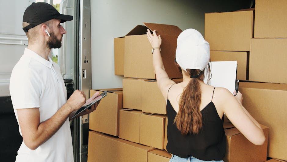A man and a woman working together during a home relocation process in a brightly lit interior space. The man, wearing a white polo shirt, black cap, and wireless earbuds, is holding a clipboard and pen, inspecting or documenting the scene. The woman, dressed in a black sleeveless top and a white headscarf, is placing or organizing open cardboard boxes stacked against a wall. The boxes vary in size and are packed with packing materials such as plastic wrap and scrunched paper. Nearby, a moving trolley loaded with additional cardboard boxes is visible, indicating an active packing or loading process. The setting suggests an organized furniture transport and packing operation, with clear focus on handling and preparing household items for a move. The environment is clean, well-lit, with a door or doorway partially visible, hinting at outdoor access for the removal vehicle, supporting efficient loading and unloading for a house removal service by [COMPANY_NAME], as noted on the page titled 'Affordable removals in Tufnell Park N19 quick quotes, TUFNELL PARK'.