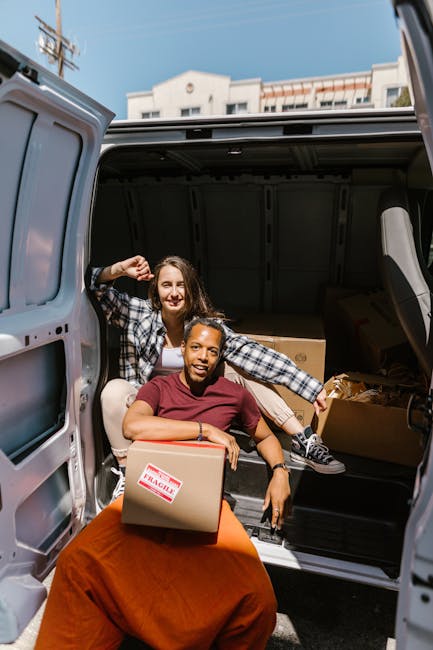 Two women are seen smiling and engaging in a home relocation activity as they pose beside a large, open van loaded with moving boxes. The woman seated at the edge of the van holds a small cardboard box marked 'FRAGILE' in red and white tape, while the woman standing behind her leans into the vehicle, gesturing with her hand. The interior of the van contains several packed cardboard boxes and wrapped items, indicating the packing stage of a furniture transport and moving process. The scene is set outdoors during daylight, with a residential building and utility poles visible in the background, suggesting the vehicle is parked near a home ready for the loading process. The image captures the careful handling of household belongings during the packing and loading phase, supported by [COMPANY_NAME]'s professional removals services documented on the page for affordable removal solutions in Tufnell Park N19.