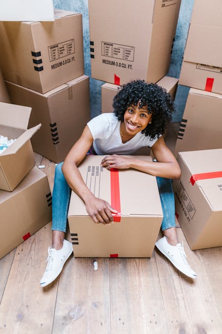 A person wearing a black T-shirt and beige pants is standing beside a woman with long, curly blonde hair, who is leaning over a cardboard box labeled 'FRAGILE'. The woman is inside a room with teal pa
