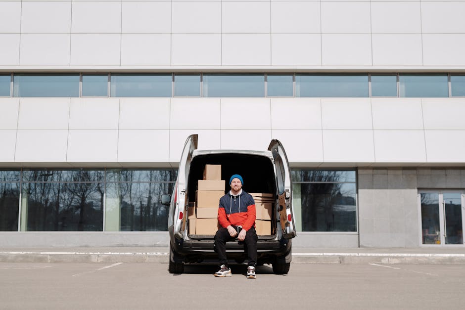 A man sits on the open back of a black cargo van parked outside a modern building with large glass windows and a grey facade. The van's interior contains several cardboard boxes stacked neatly, ready for home relocation. The boxes vary in size and are secured with plastic wrap to prevent shifting during transportation. The man is dressed in casual clothing, including a blue beanie, a red and navy jacket, black pants, and sneakers, and is smiling slightly at the camera. The loading process appears to be in progress, with the van positioned on a paved area directly outside an entrance, suggesting ongoing furniture transport and packing for a house move, supported by the related services of the removals company, [COMPANY_NAME]. The cloudy sky and leafless trees reflected in the windows indicate an outdoor setting possibly during late autumn or winter, emphasizing the logistical effort involved in a swift same-day move.
